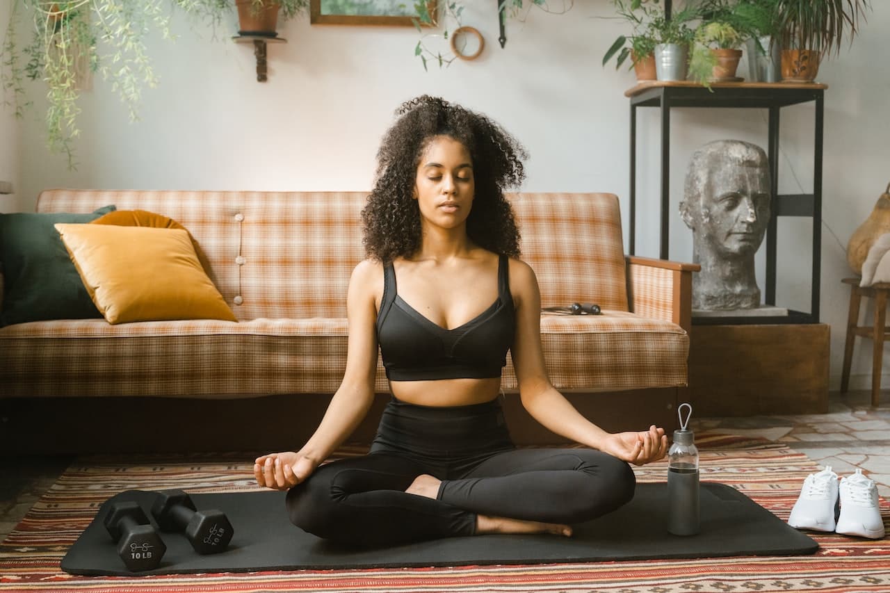 A person doing yoga, sitting cross-legged with eyes closed, in a peaceful and relaxed state. The person appears to be in a meditative state, with arms and legs in different positions, and a faint smile on their lips. The image suggests that the person has used medical cannabis before starting their yoga practice, as it promotes relaxation and helps to induce a peaceful state of mind. The background shows a calm and serene environment, with soft lighting and a natural landscape visible through a large window.
